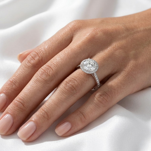 Close-up of a round brilliant diamond halo engagement ring with a pavé band worn on a finger.