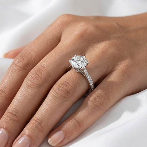 Close-up of a round brilliant diamond engagement ring with a pavé band worn on a finger.